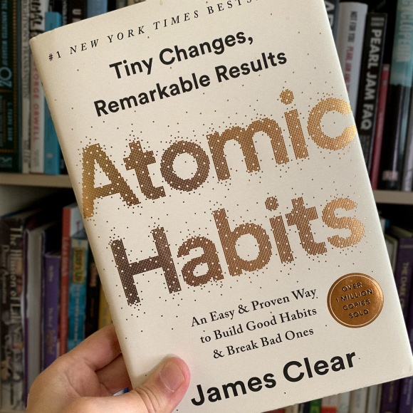 A photo of James Clear’s book ‘Atomic Habits’ being held up in front of a bookshelf.